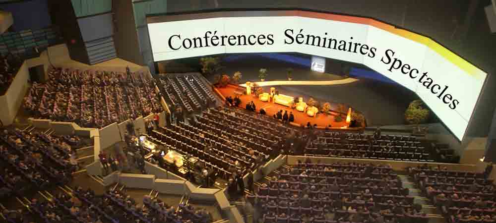 Grande salle de conférence remplie de spectateurs, scène éclairée avec orateurs, écran panoramique affichant 'Conférences Séminaires Spectacles'.
